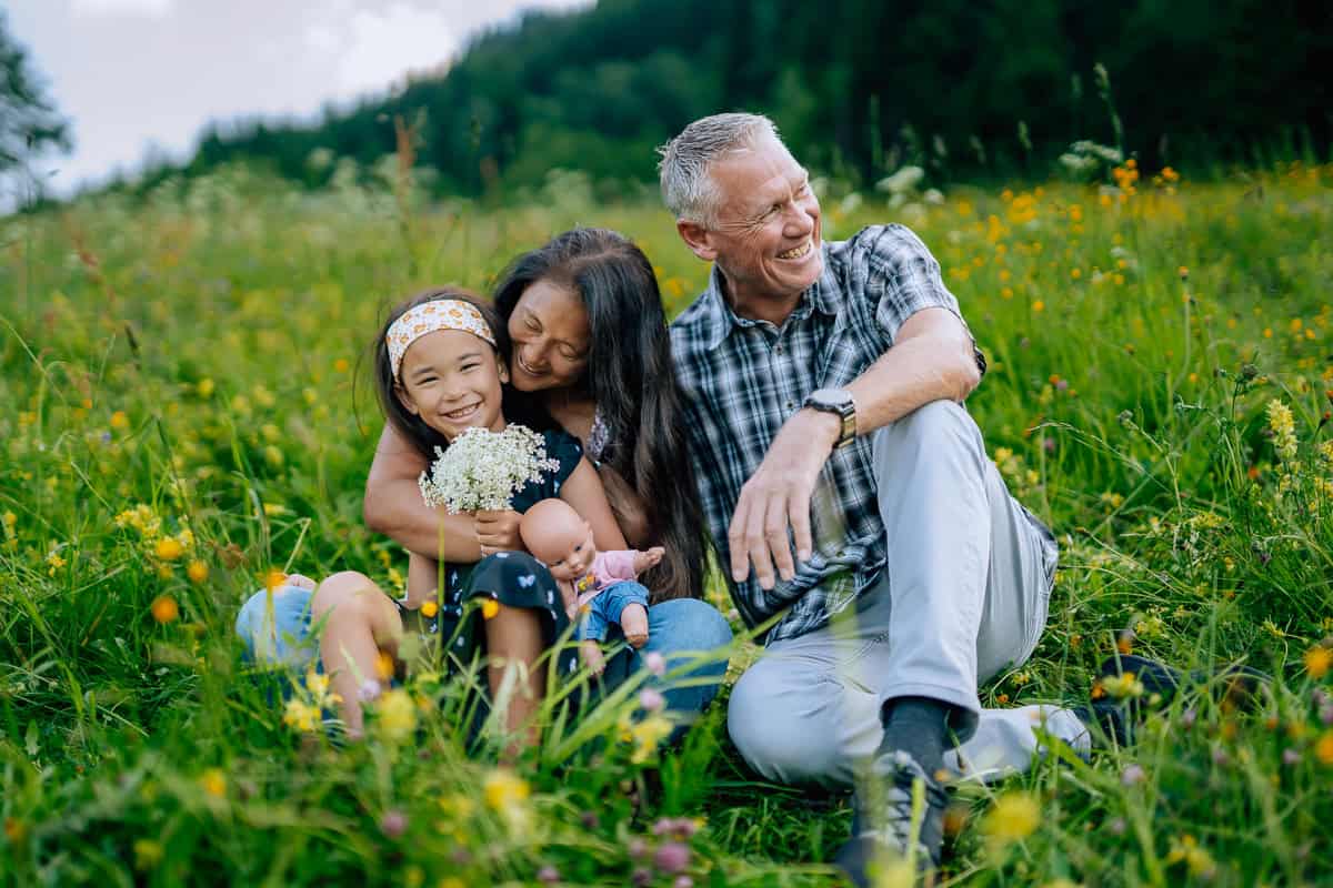 A joyful multigenerational family, including a young girl, a woman, and an elderly man, happily sitting in a vibrant flower field, embracing and laughing during their worldschooling journey experience.