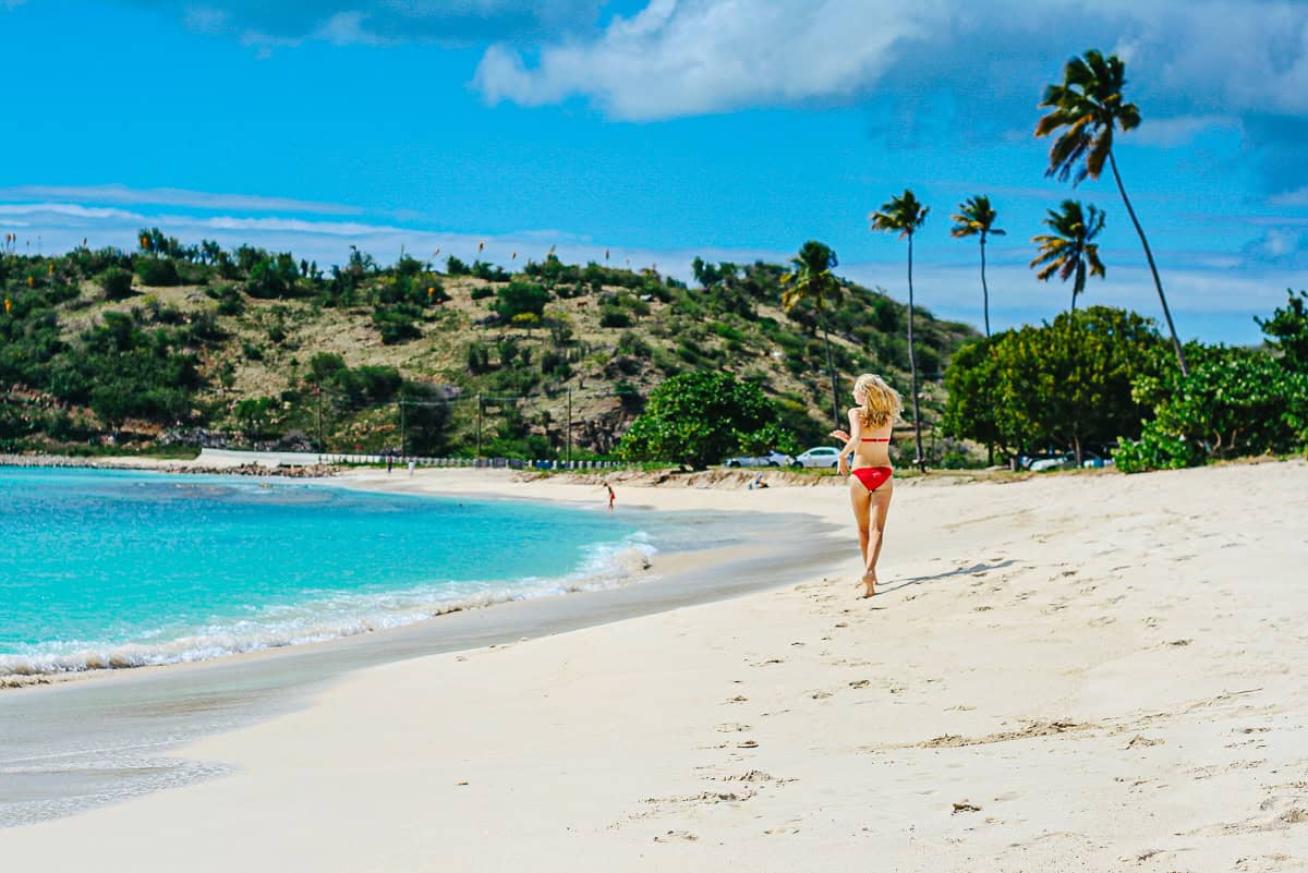 A woman in a red bikini walks along a beautiful sandy beach with turquoise waters, lush greenery, and palm trees, representing the ideal setting for worldschooling journeys and adventurous global learning experiences.